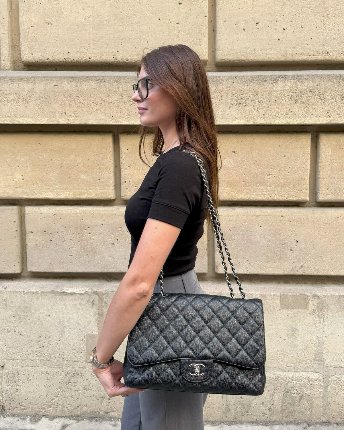 Sac Chanel noir matelassé porté à l’épaule devant une façade parisienne en pierre.